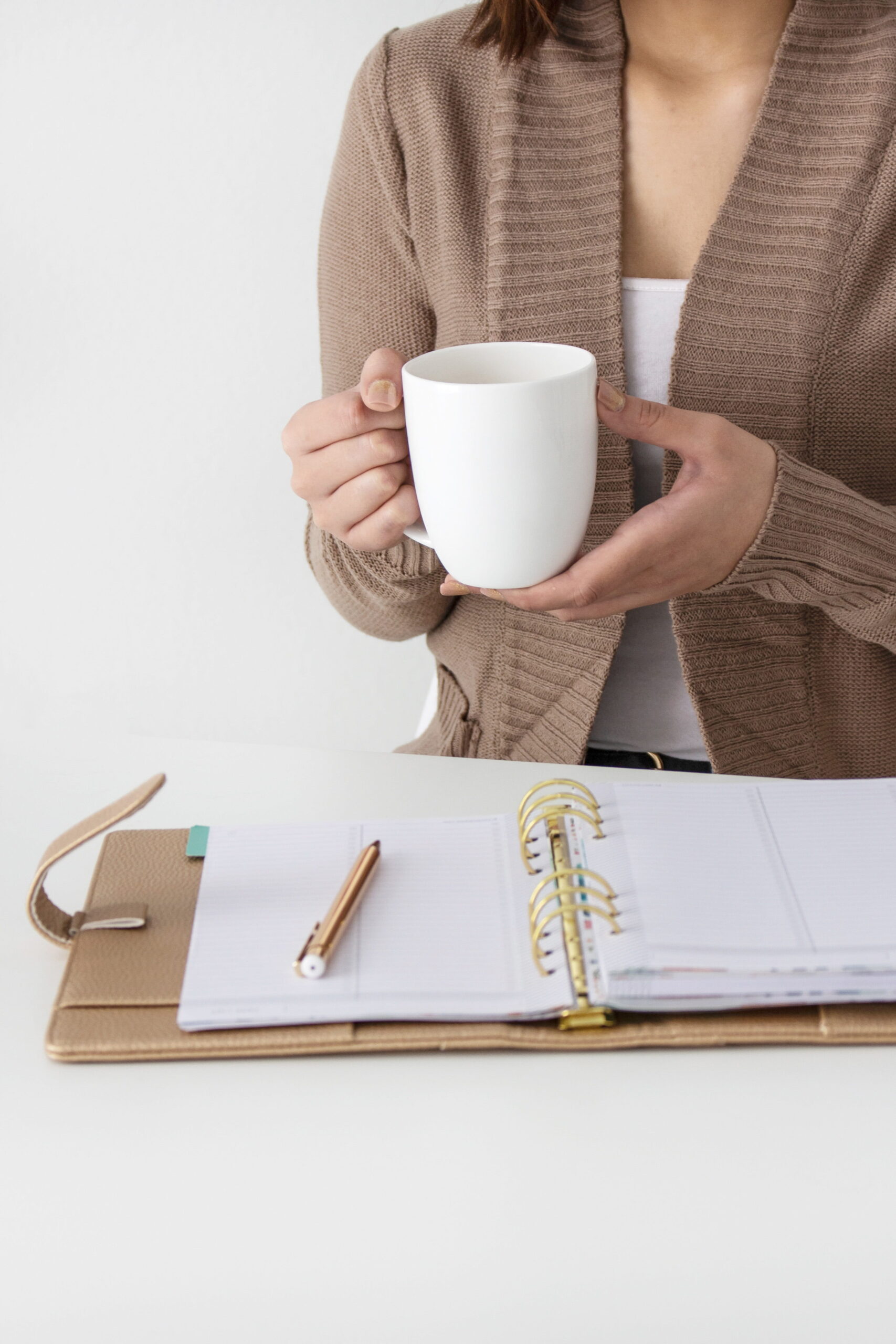 A virtual assistant in a beige cardigan holds a white mug. In front of her is an open planner with a pen, set on a white table, conveying a calm, organized atmosphere.