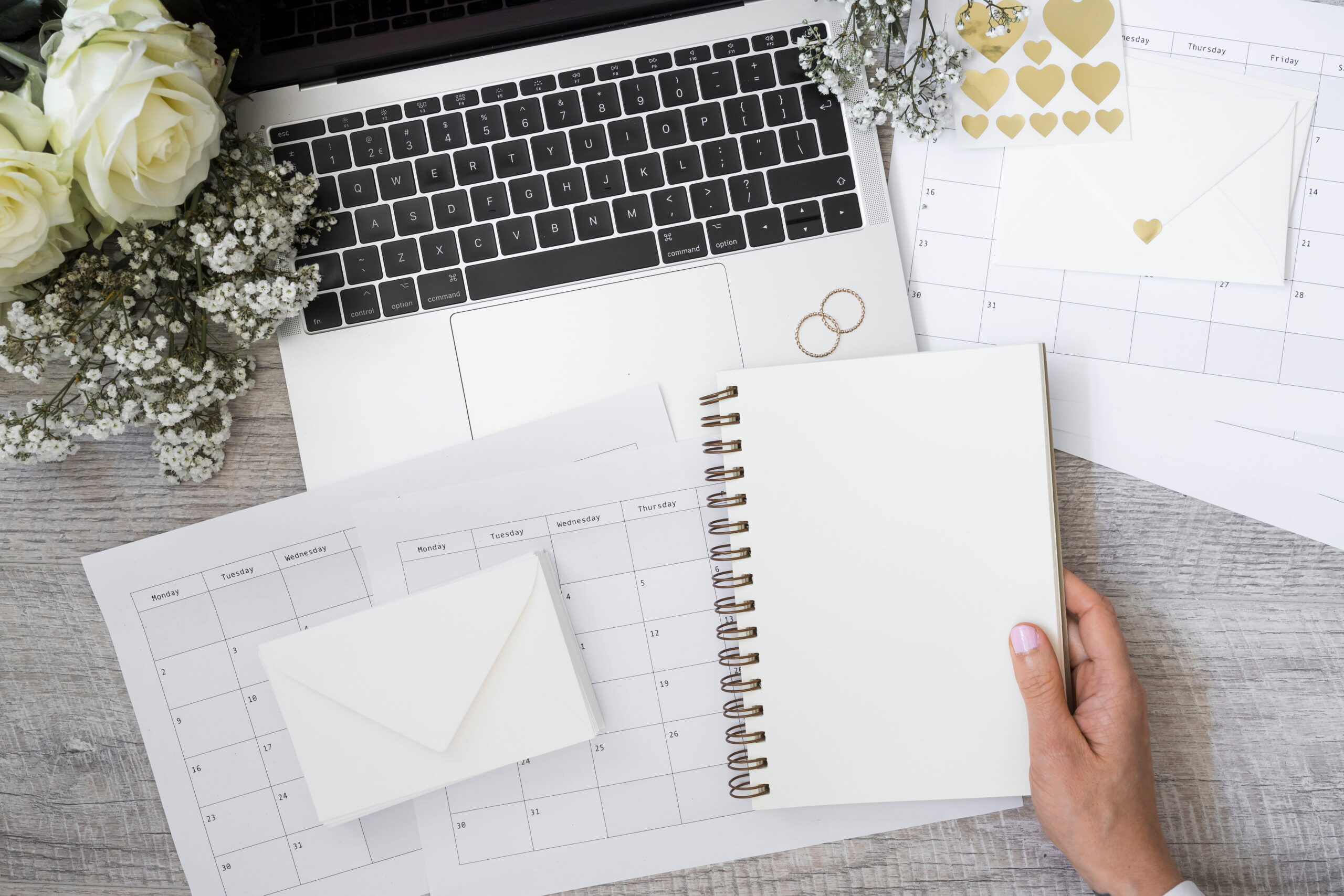 A virtual assistant holds a blank notebook over a desk with a laptop, planner, and envelopes. White roses and heart stickers convey a theme of planning.