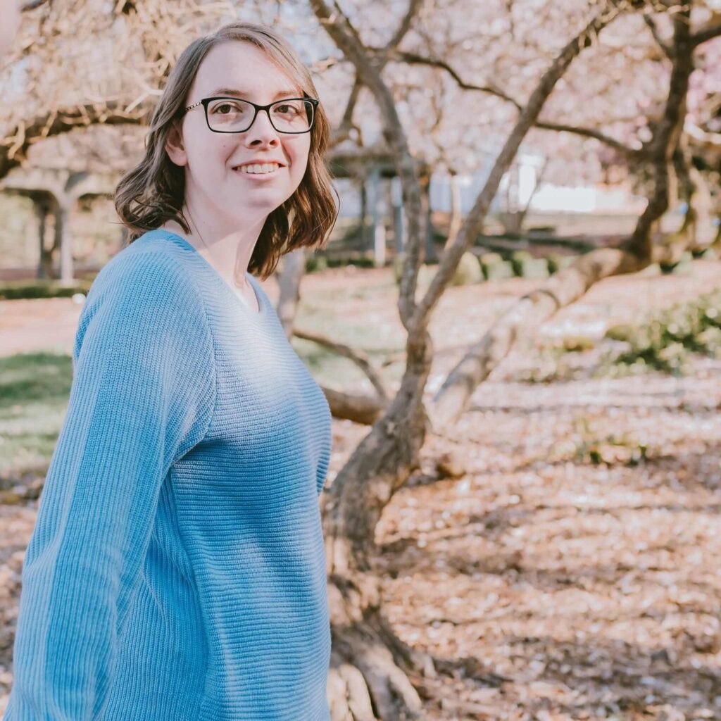 Kristin Gunner, a virtual assistant for small business owners, smiling in glasses and a blue sweater, standing in a sunlit park with leafless trees and soft, warm lighting, creating a relaxed, cheerful mood.