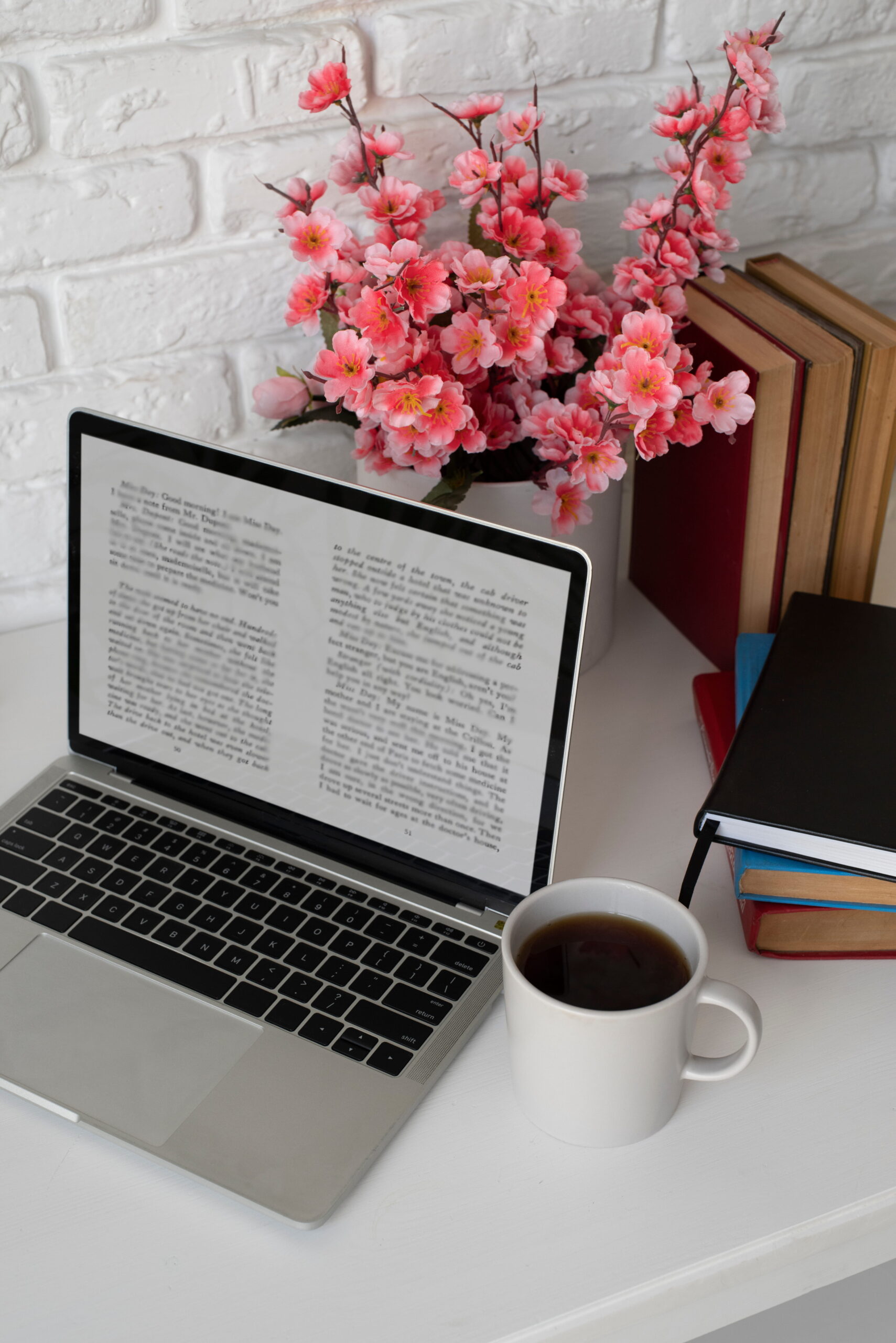 A laptop displaying text sits on a white desk next to a cup of coffee, pink cherry blossoms, and stacked books, creating a cozy and productive atmosphere.