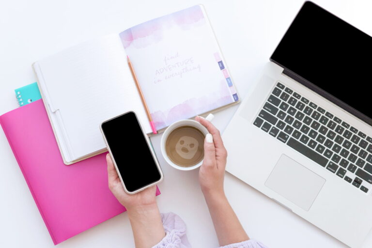 Woman's hands holding a coffee cup and smartphone on a white desk with a laptop and notebook nearby.