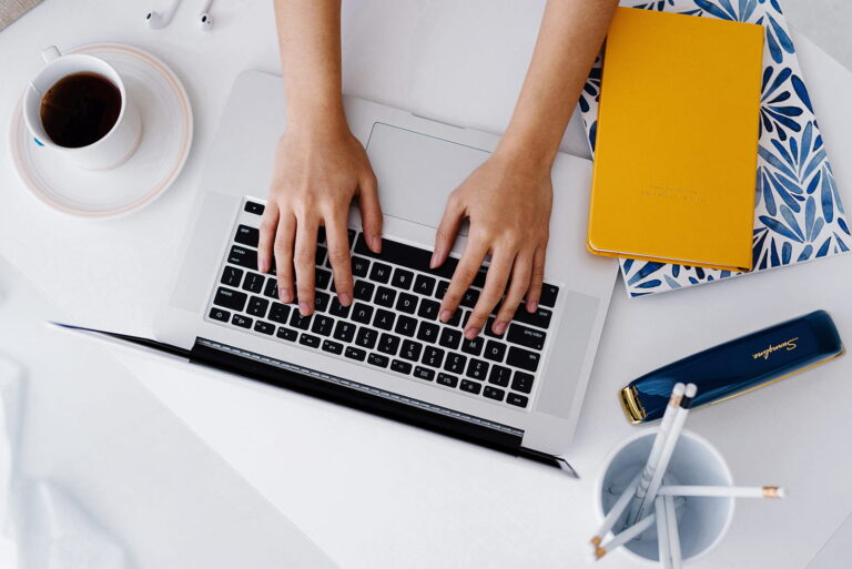 A woman's hands typing on a laptop, with a coffee cup and a notebook beside it.