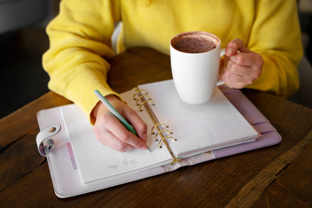 Woman writing in a notebook while sipping coffee, surrounded by a cozy workspace.