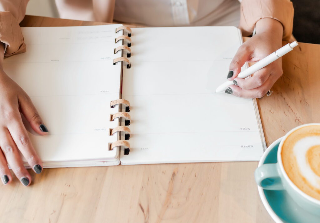 Virtual assistant holding a pen, writing in a planner on a wooden table, with a cup nearby.