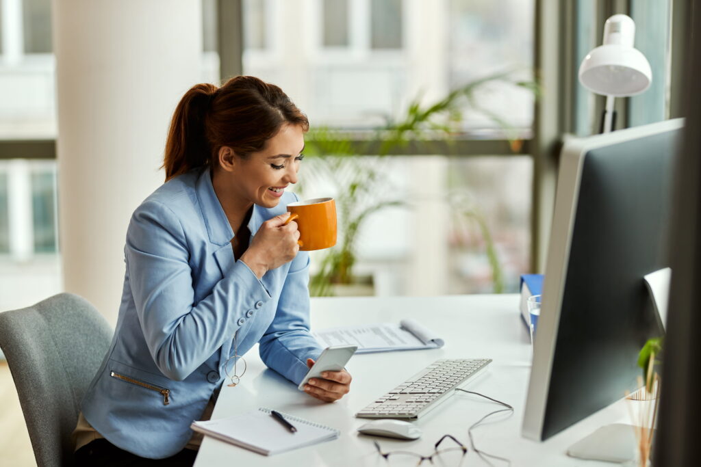 A woman sits at a desk, enjoying a cup of coffee while working on her laptop.