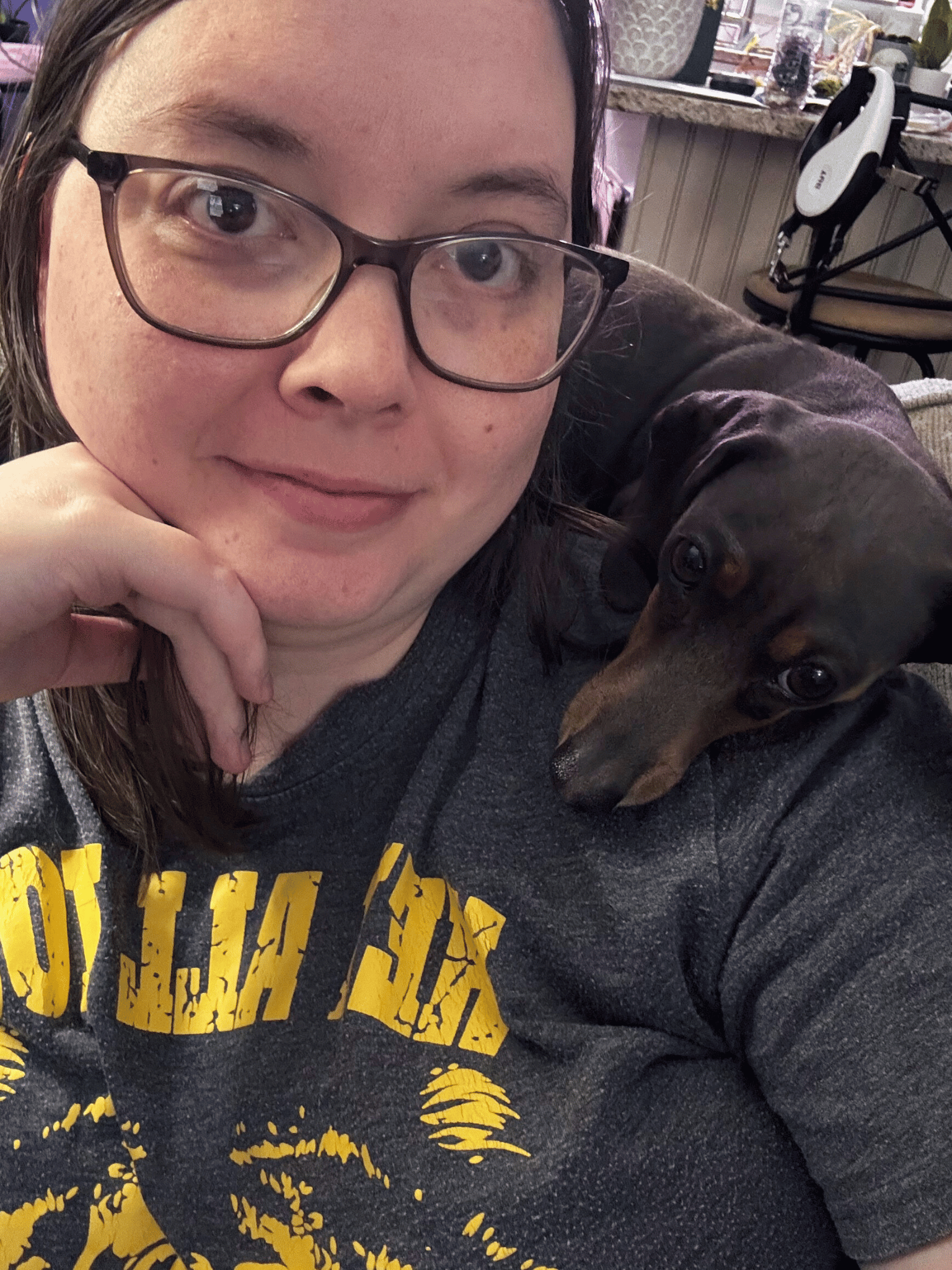 A woman in glasses and a yellow shirt smiles with a dachshund puppy on her shoulder.